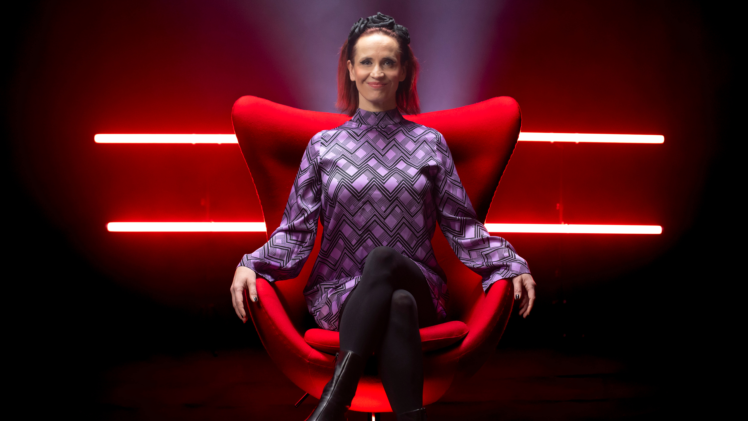 Maija Vilkkumaa istuu punaisella tuolilla pimeässä huoneessa ja hymyilee. Takana neonvalot. 